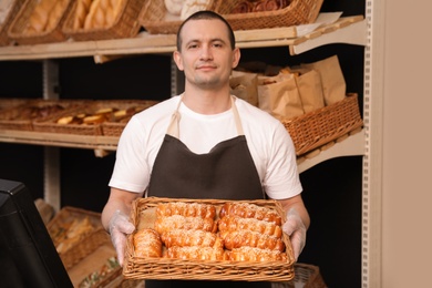 Photo of Portrait of professional baker holding tray with fresh buns near showcase in store