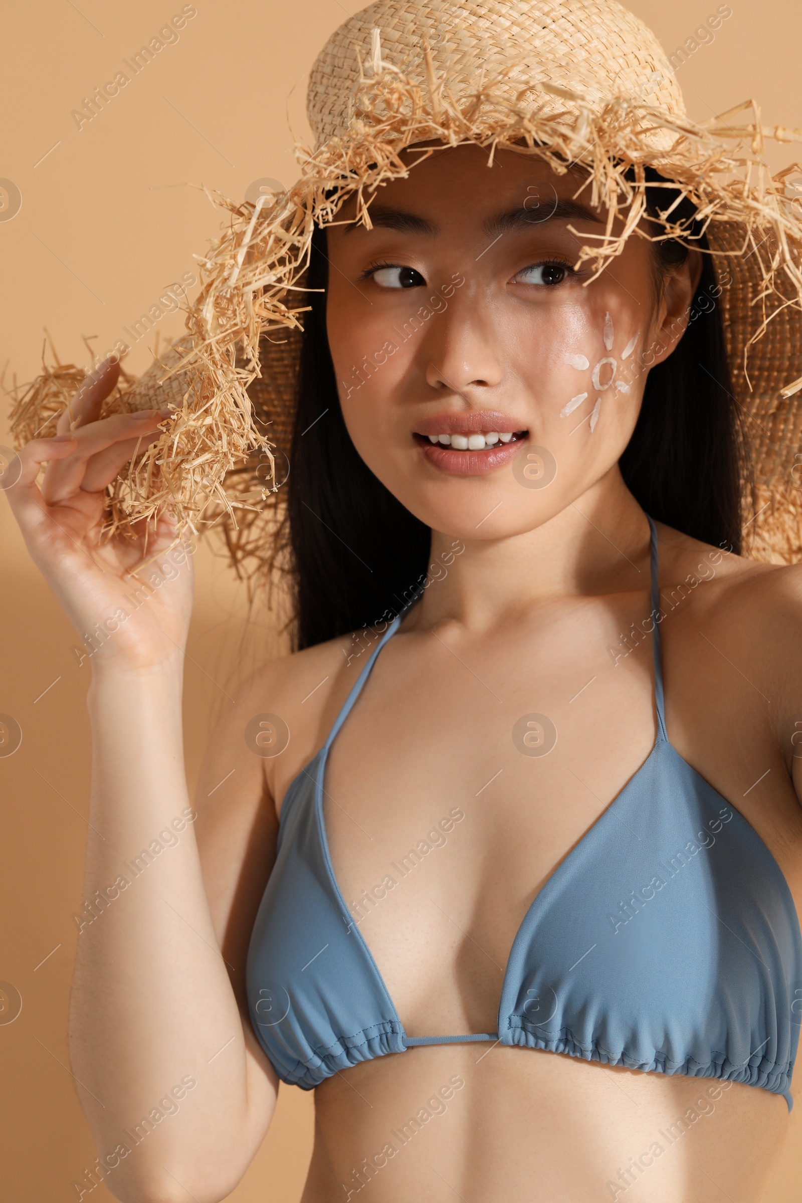 Photo of Beautiful young woman in straw hat with sun protection cream on her face against beige background