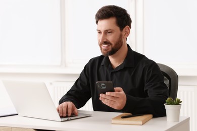 Photo of Smiling man with smartphone using laptop at table in office