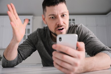 Photo of Emotional man with smartphone at table in kitchen. Online hate concept