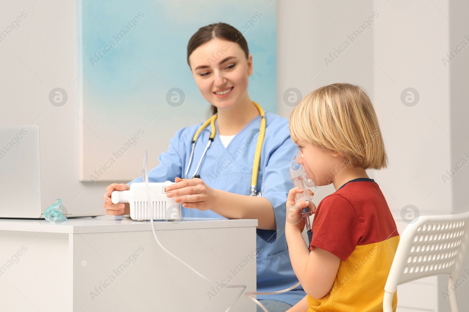 Photo of Medical assistant sitting near sick little boy while he using nebulizer for inhalation in hospital