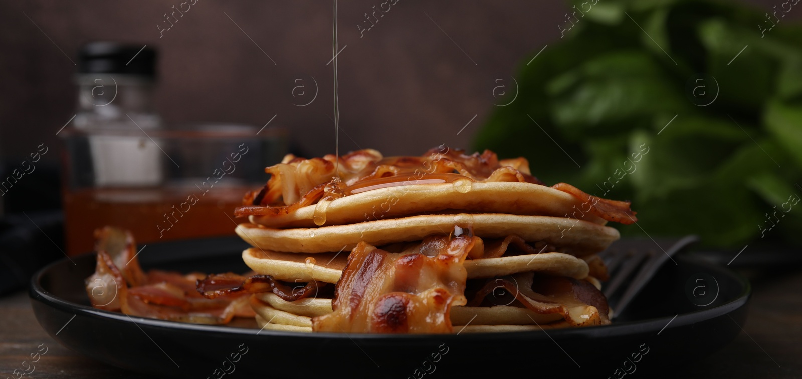 Photo of Pouring maple syrup onto delicious pancakes with fried bacon on wooden table, closeup