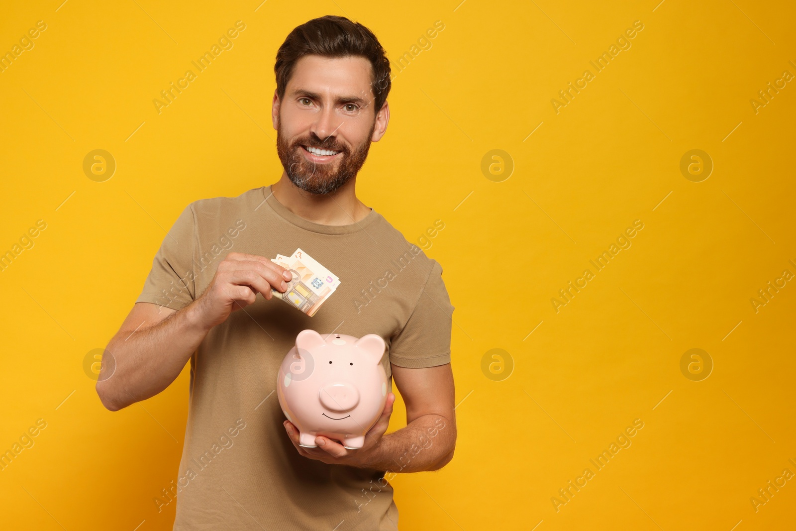Photo of Happy man putting money into piggy bank on orange background, space for text
