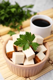 Photo of Bowl of smoked tofu cubes, soy sauce and parsley on bamboo mat, closeup