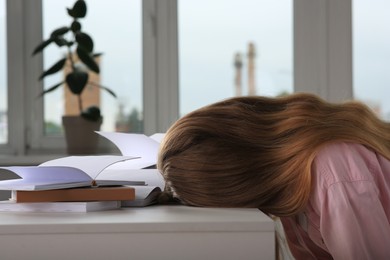 Photo of Young tired woman sleeping near books at white table in room