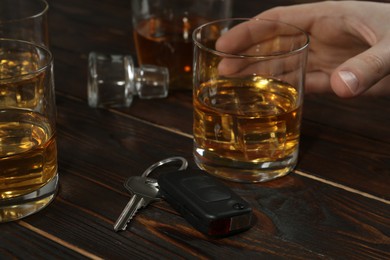 Photo of Man holding glass of alcohol near car key at wooden table, closeup. Dangerous drinking and driving