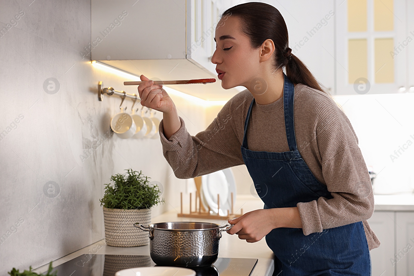 Photo of Smiling woman with wooden spoon tasting tomato soup in kitchen