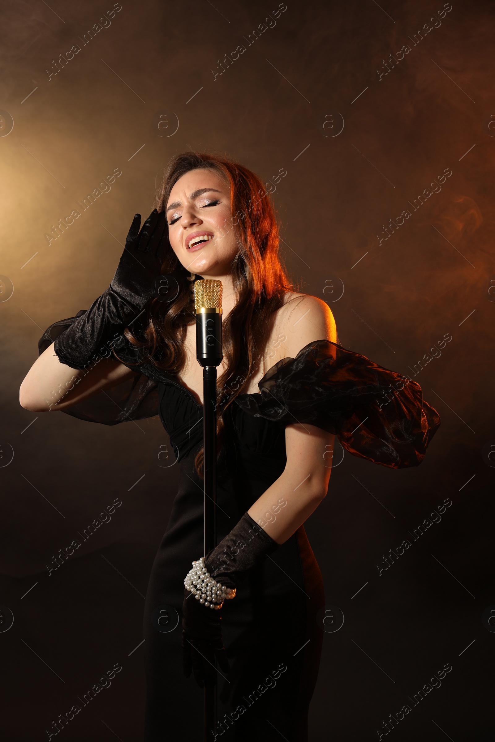 Photo of Beautiful young woman with microphone singing on dark background with smoke