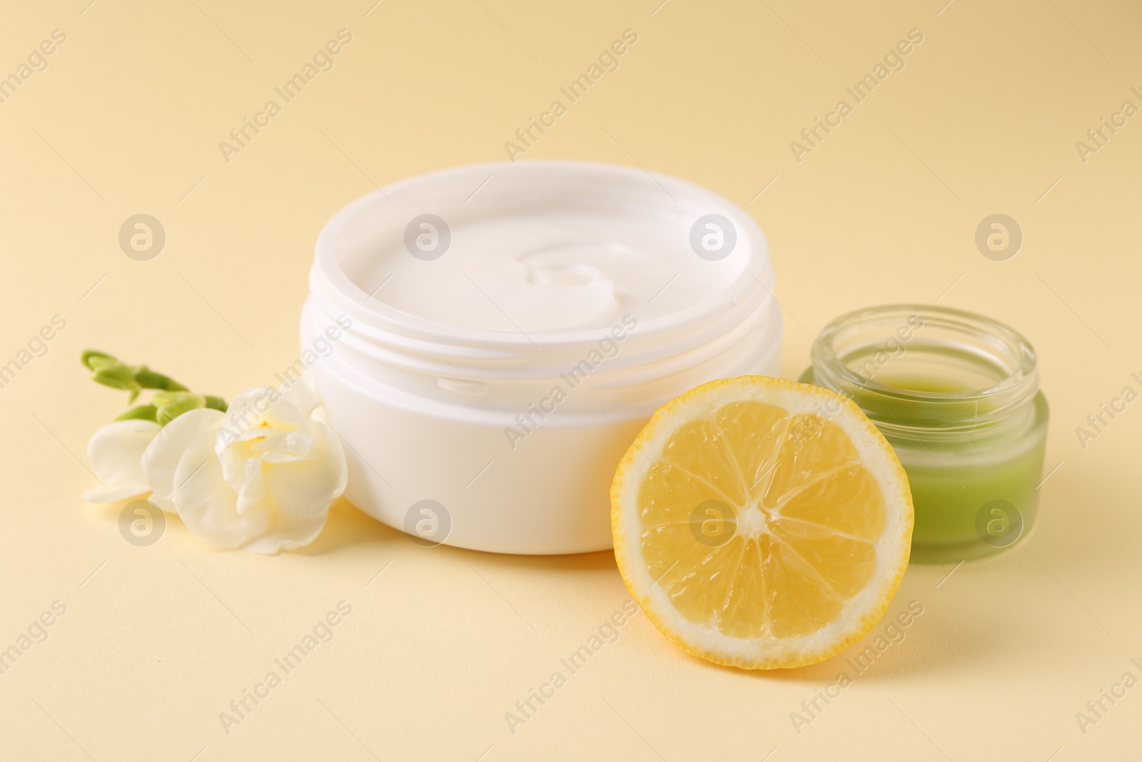 Photo of Moisturizing cream in open jars, lemon and freesia flower on beige background. Body care products