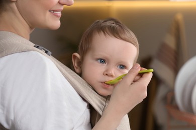 Mother feeding her cute child in sling (baby carrier) indoors