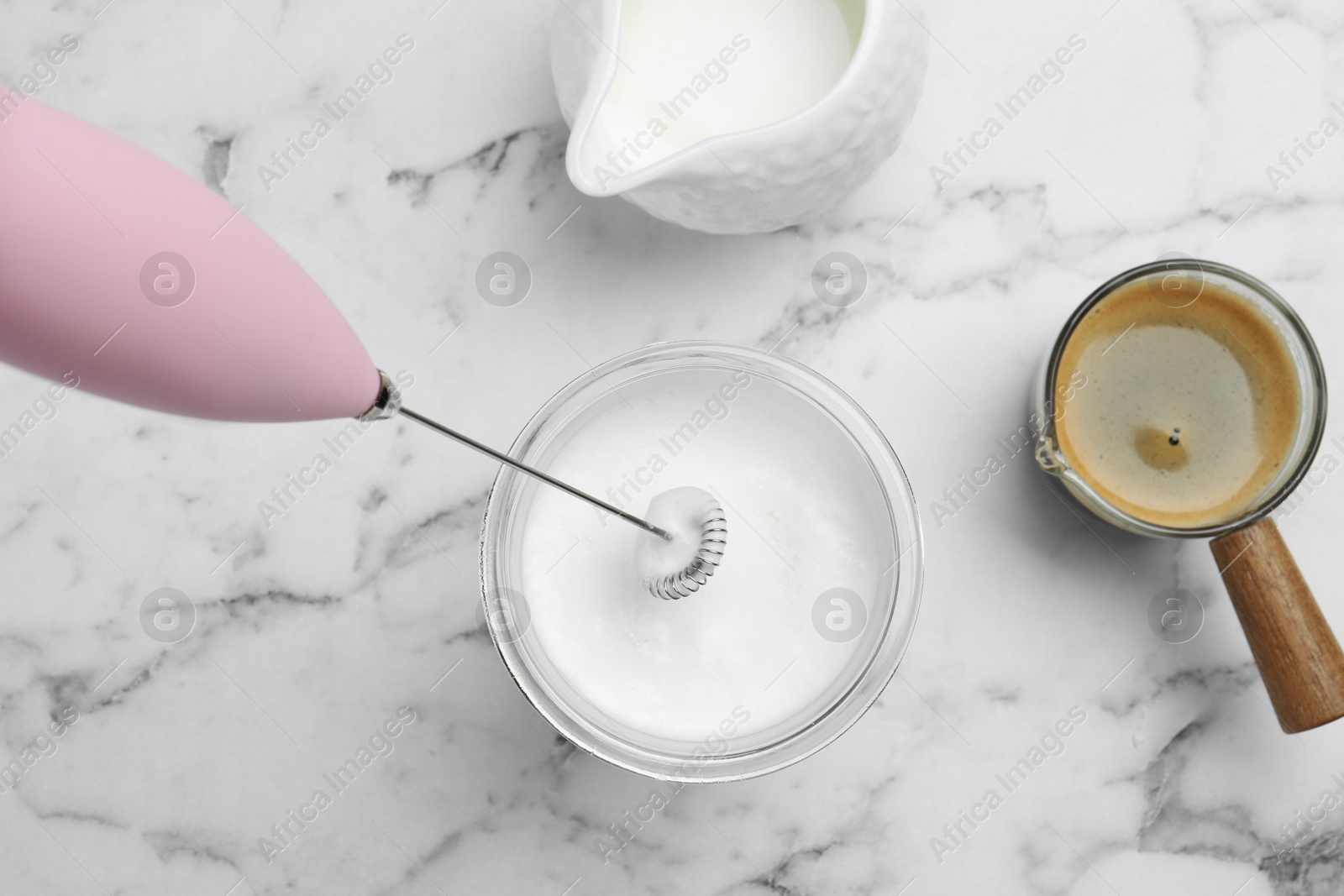 Photo of Whisking milk in glass with mini mixer (milk frother) at white marble table, flat lay