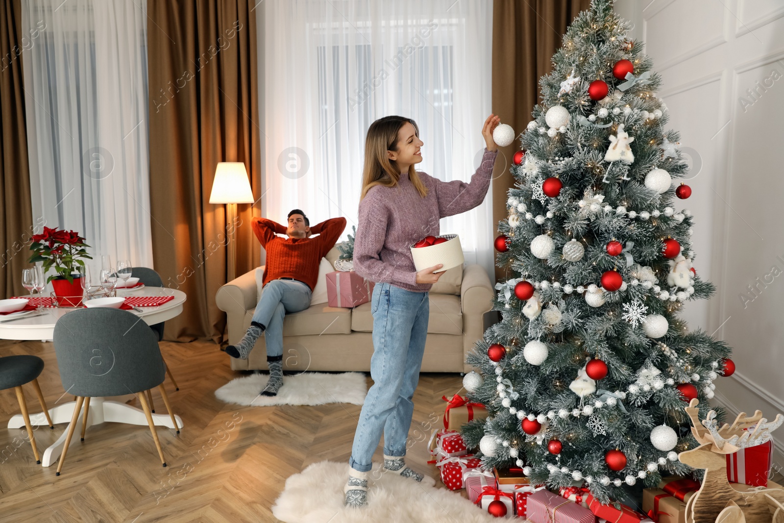 Photo of Young woman decorating Christmas tree while her boyfriend resting on sofa at home