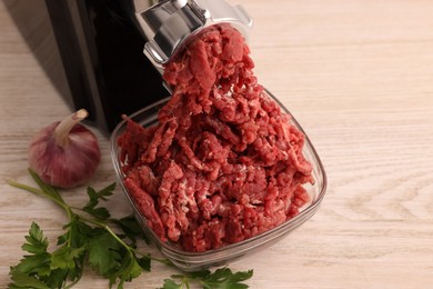 Photo of Electric meat grinder with beef mince, garlic and parsley on white wooden table, space for text