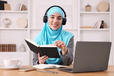 Photo of Muslim woman in headphones studying near laptop at wooden table in room