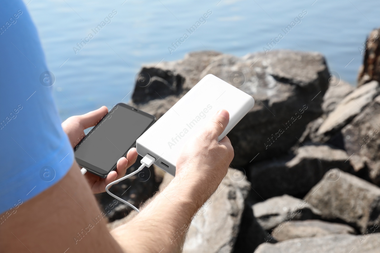 Photo of Man charging mobile phone with power bank on rocky mountain near river, closeup