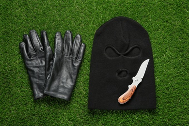 Black knitted balaclava, gloves and knife on green grass, flat lay