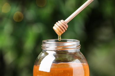 Photo of Dripping tasty honey from dipper into jar on green blurred background