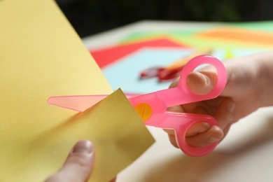 Photo of Child cutting color paper with plastic scissors at table, closeup