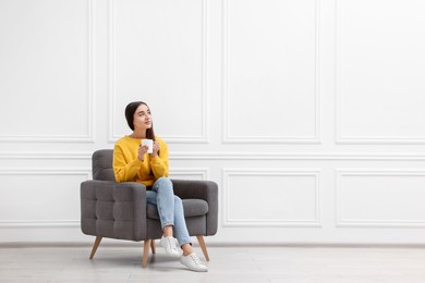 Photo of Beautiful woman with cup of drink sitting in armchair near white wall indoors, space for text