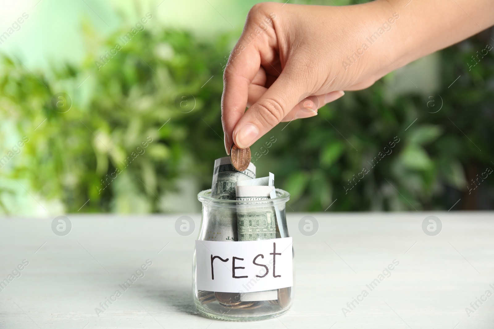 Photo of Woman putting coin into jar with tag REST on white wooden table, closeup