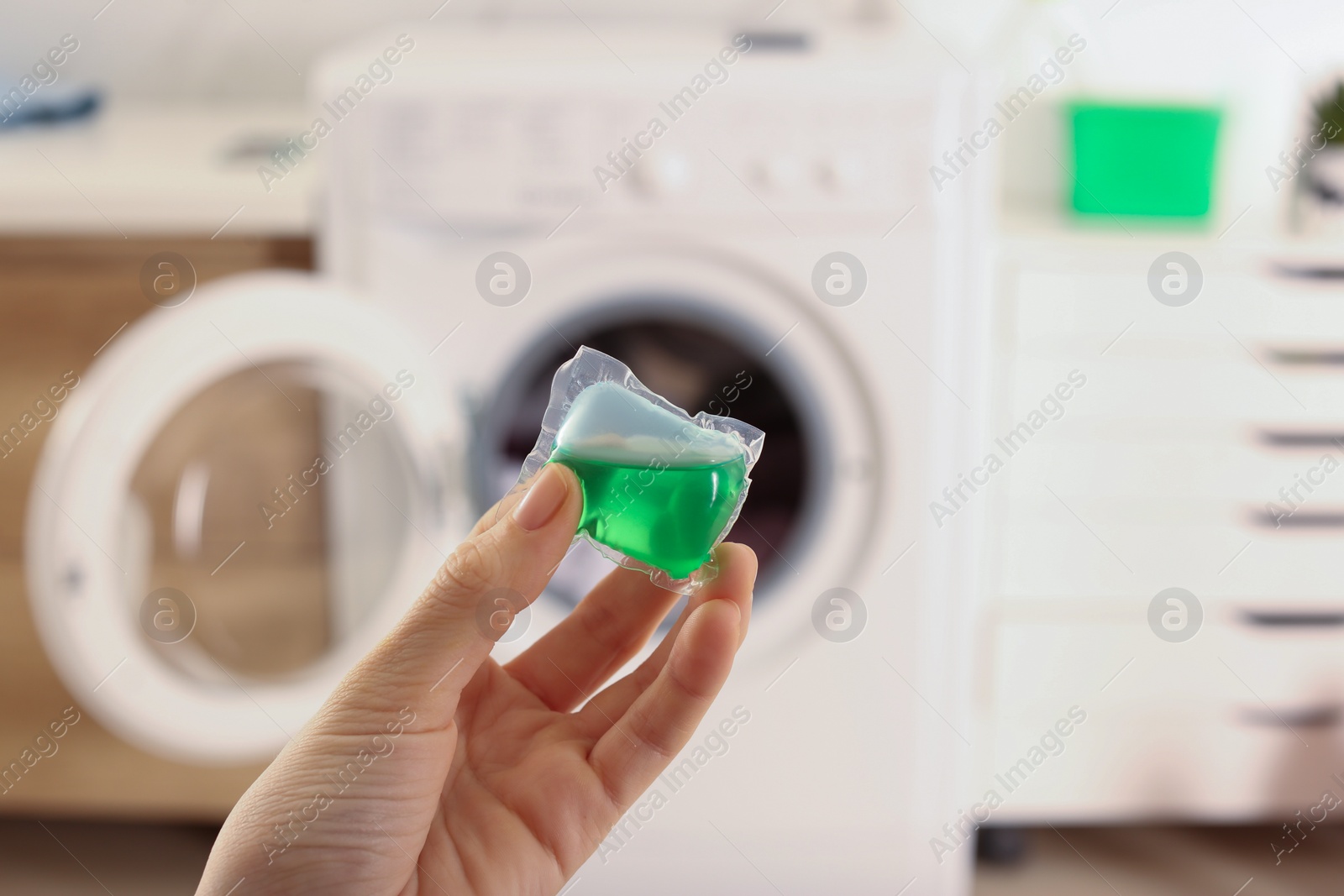 Photo of Woman holding laundry detergent capsule near washing machine indoors, closeup