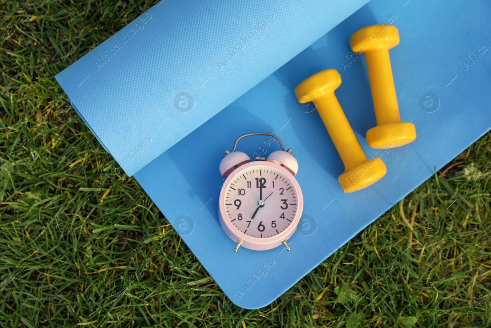 Photo of Alarm clock, dumbbells and fitness mat on green grass, flat lay. Morning exercise