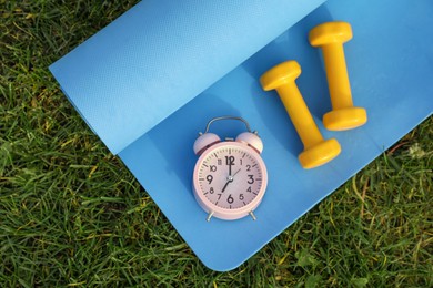 Photo of Alarm clock, dumbbells and fitness mat on green grass, flat lay. Morning exercise