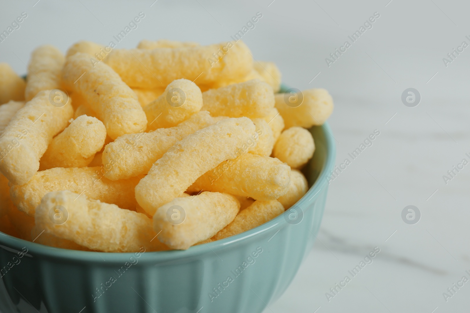 Photo of Bowl of delicious crispy corn sticks on white marble table, closeup