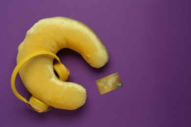 Photo of Yellow travel pillow, headphones and credit card on purple background, flat lay. Space for text