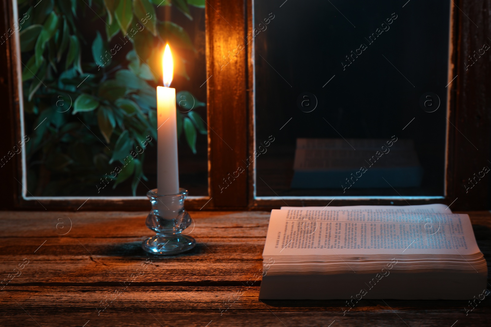 Photo of Burning candle and Bible on wooden table near window at night