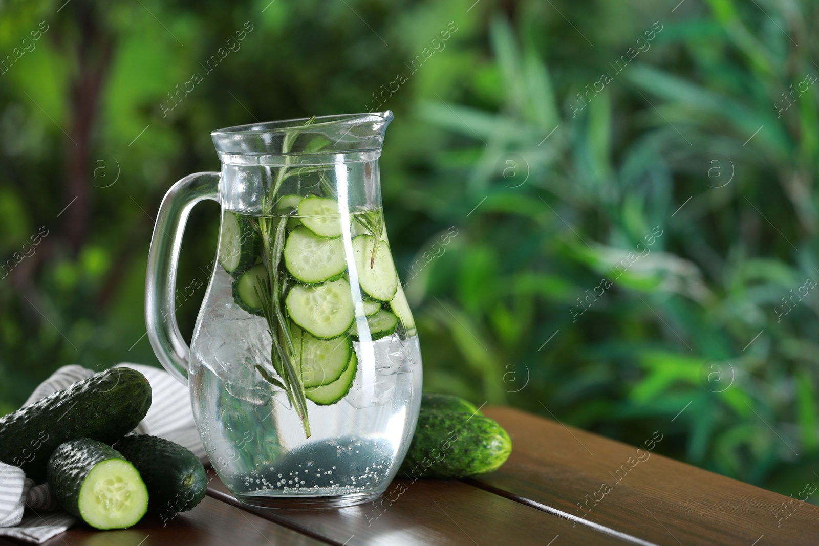 Photo of Refreshing cucumber water with rosemary in jug and vegetables on wooden table against blurred green background. Space for text