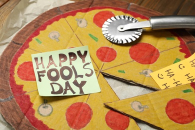 Photo of Cardboard pizza and Happy Fools' Day note on wooden table, closeup