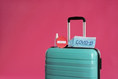 Antiseptic spray, stop sign and protective masks with inscription COVID-19 on suitcase against pink background, space for text. Travel restriction during coronavirus pandemic