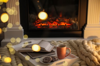 Photo of Cup of coffee, cookies and books on knitted blanket near burning fireplace indoors. Cozy atmosphere