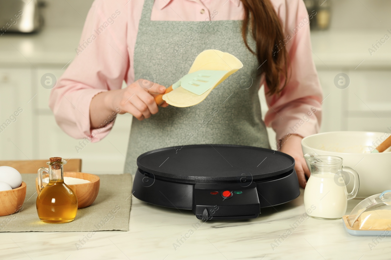 Photo of Woman cooking delicious crepe on electric maker at white marble table in kitchen, closeup