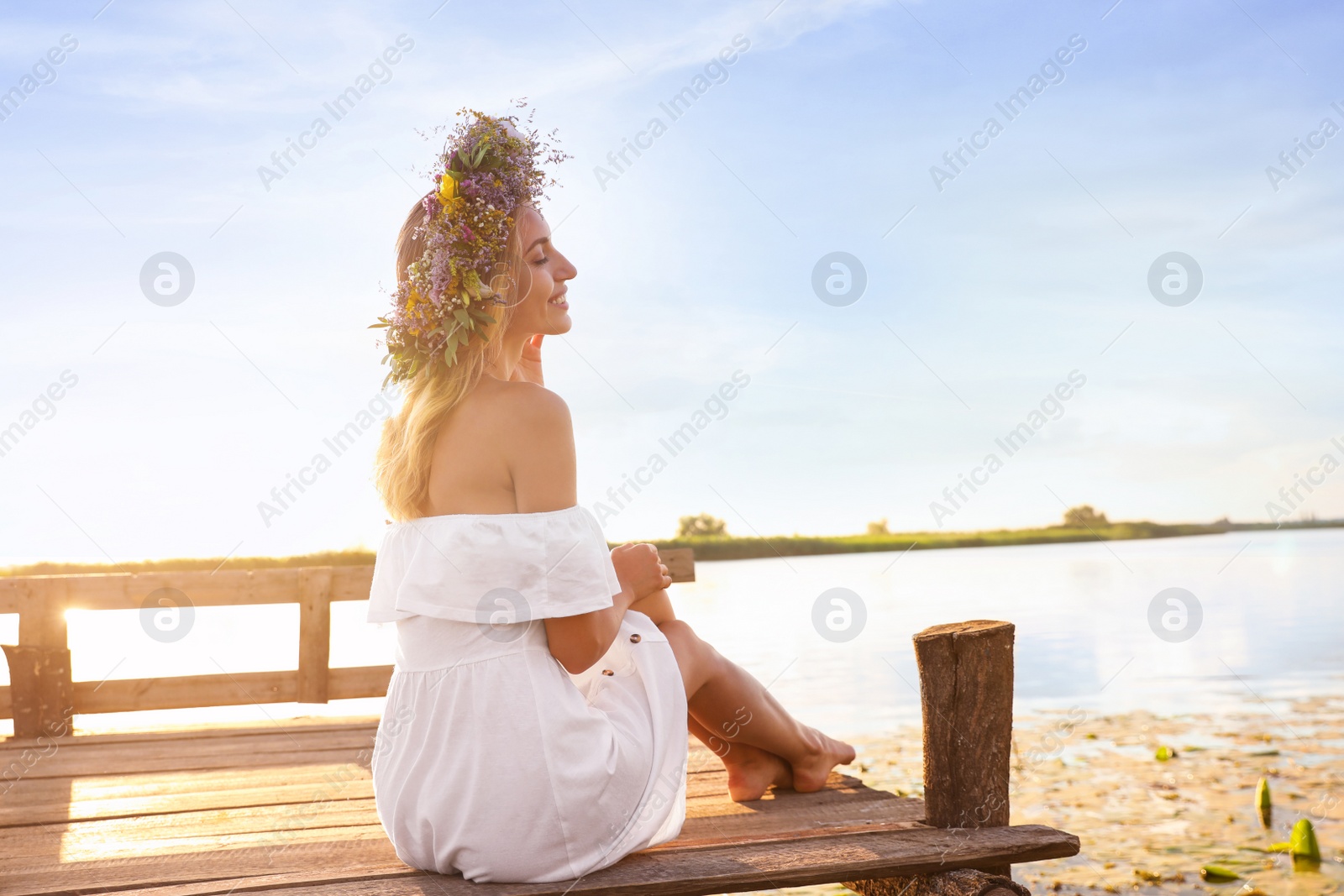 Photo of Young woman wearing wreath made of beautiful flowers on pier near river