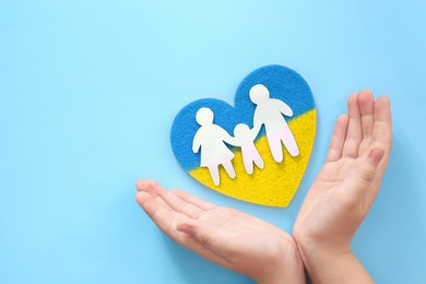 Stop war in Ukraine. Child holding hands near heart with colors of Ukrainian flags and paper family on light blue background, top view