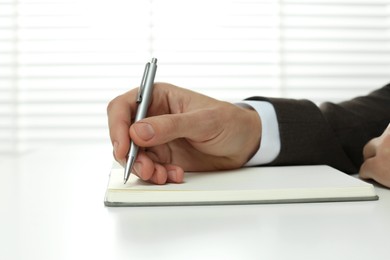 Photo of Man writing in notebook at white table indoors, closeup