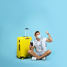 Male tourist in protective mask holding passport with ticket and suitcase on turquoise background, space for text