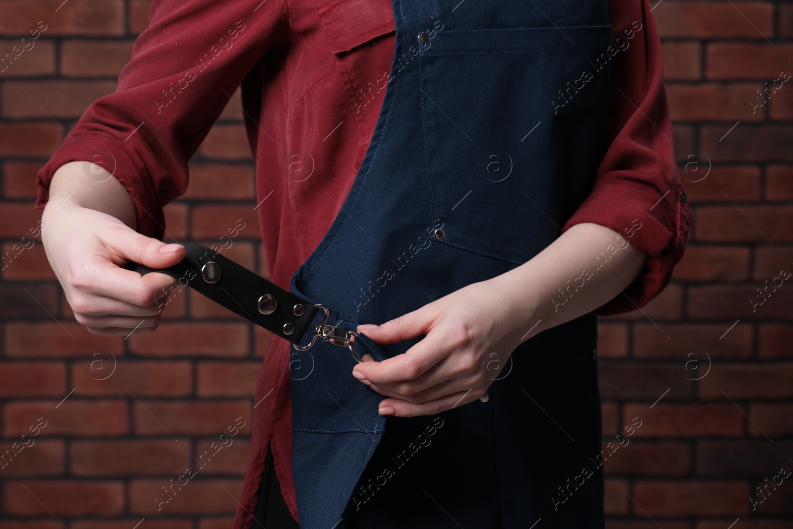 Photo of Woman putting on blue apron near red brick wall, closeup