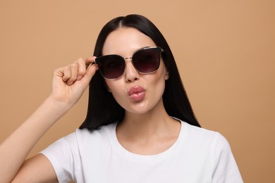 Beautiful young woman in stylish sunglasses blowing kiss on beige background