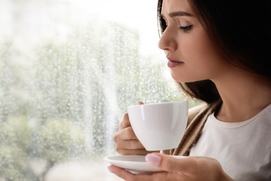 Thoughtful beautiful woman with cup of coffee near window indoors on rainy day