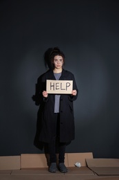 Photo of Poor young woman with HELP sign standing near dark wall