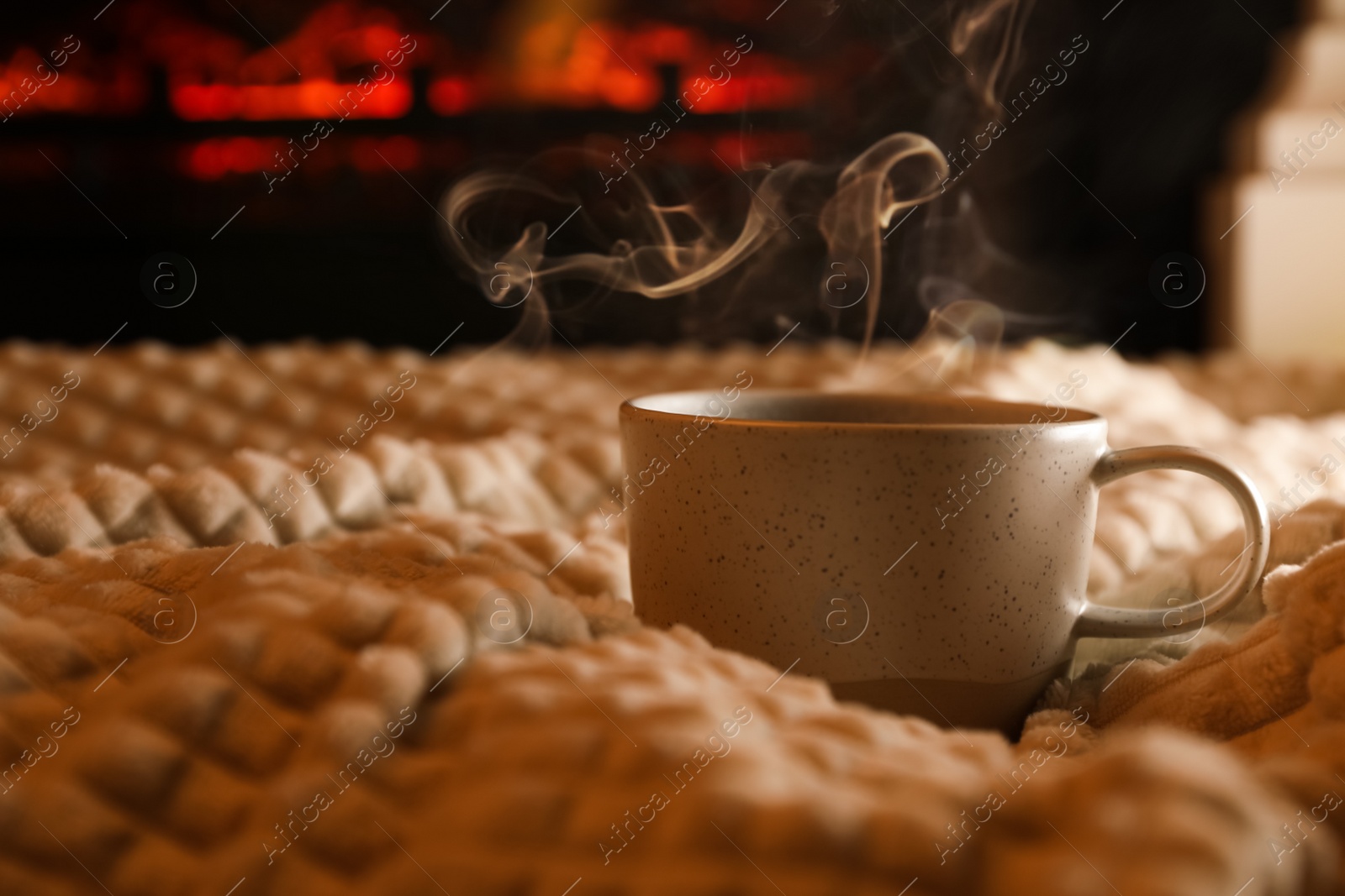 Photo of Cup of hot drink on knitted plaid near fireplace indoors. Space for text