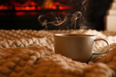 Cup of hot drink on knitted plaid near fireplace indoors. Space for text