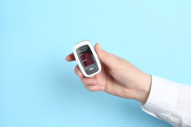 Woman holding modern fingertip pulse oximeter on light blue background, closeup