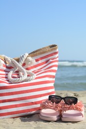 Photo of Stylish striped bag with slippers and sunglasses on sandy beach near sea