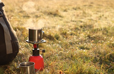 Camping burner with mug of hot drink near backpack on grass outdoors. Space for text