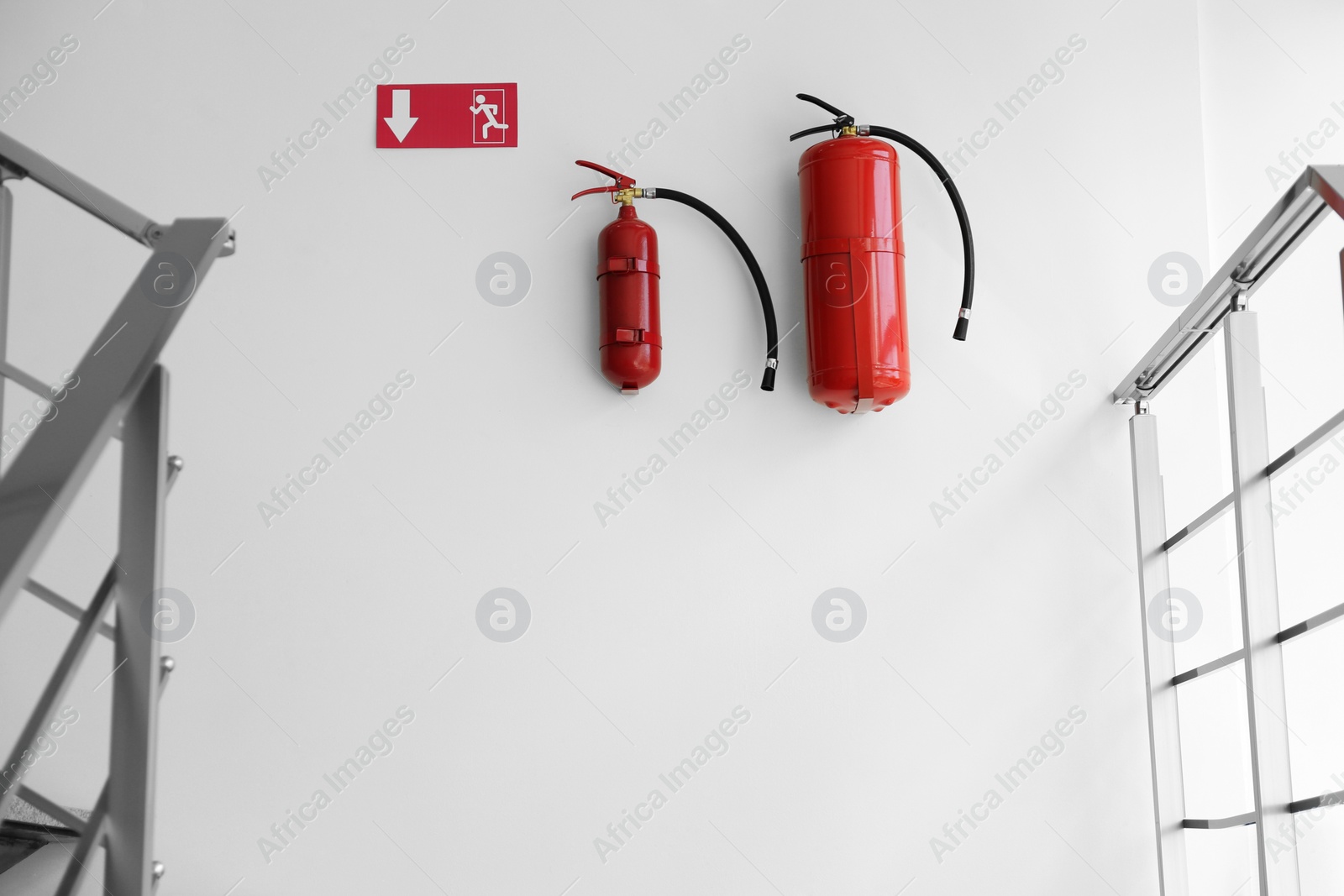 Photo of Fire extinguishers and emergency exit sign on white wall near staircase indoors