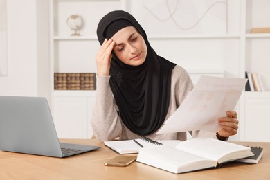 Tired Muslim woman in hijab working near laptop at wooden table in room